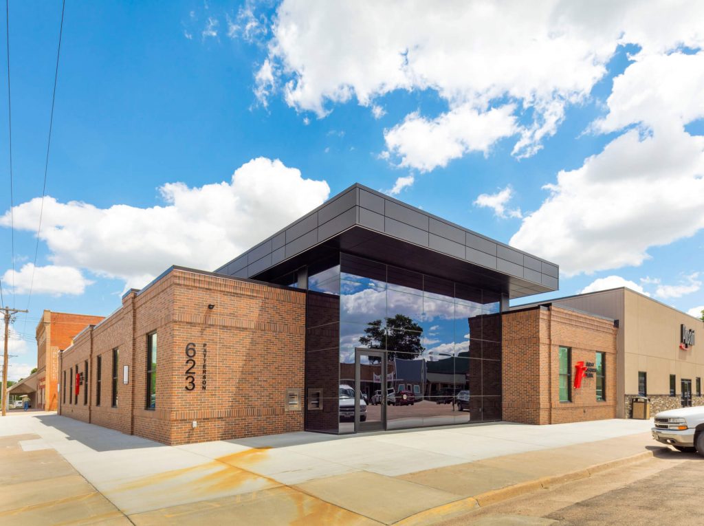 Corner view of facade of new, brick bank with large, tinted window entrance under a blue sky with large, white, puffy clouds