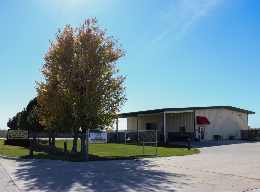 Cream colored, industrial building next to a row of mid-sized trees and a large concrete parking lot