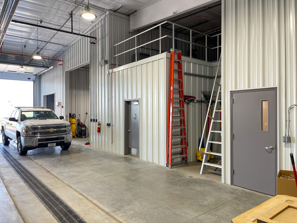 Inside an industrial building with metal walls and a white pickup truck, as well as an orange ladder and a white ladder that leads up to a loft